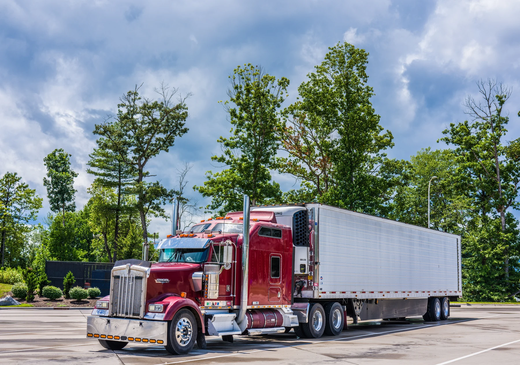 Red American semi truck with a white trailer parked beside green trees